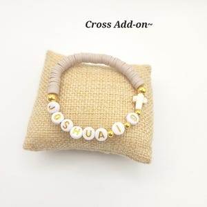 May include: A bracelet featuring beige clay heishi beads, white letter beads spelling JOSHUA 19, gold-tone spacer beads, and a small white cross charm. The text Cross Add-on~ is visible above the bracelet, which is displayed on a square burlap pillow.
