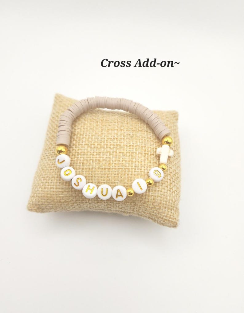 May include: A bracelet featuring beige clay heishi beads, white letter beads spelling JOSHUA 19, gold-tone spacer beads, and a small white cross charm. The text Cross Add-on~ is visible above the bracelet, which is displayed on a square burlap pillow.