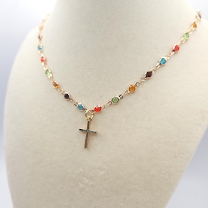 May include: Gold-toned necklace with a delicate chain, featuring colorful gemstone beads and a small cross pendant. The necklace is displayed on a mannequin, showcasing its design and details. The gemstones are in various colors.