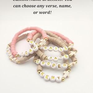 May include: A collection of beaded bracelets featuring pink and beige clay discs, gold-tone spacer beads, and white letter beads spelling names like GAGE, MICAH, GAYLE, JASE, COOPER, and KENT, with small white cross charms and the text Custom Name Bracelets!