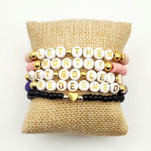 May include: Stack of beaded bracelets with white letter beads spelling "LET THEM WORTHY LET GO REDEEMED". The bracelets are in shades of pink, gold, black, and purple. A gold heart charm is on the bottom bracelet.