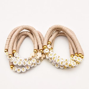 May include: Two stacks of beaded bracelets made with beige clay disc beads, gold-toned metallic spacer beads, and white round letter beads. One stack spells CHOSEN and features a small cross charm, while the other spells PETER 12.
