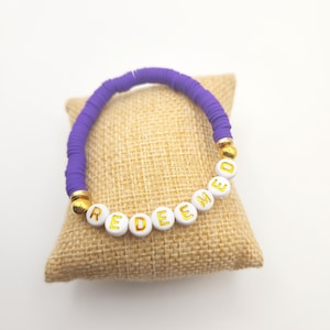 May include: A bracelet featuring purple disc-shaped beads and white spherical beads with the gold-colored text "REDEEMED" printed on them. The bracelet includes two gold-colored spherical accent beads positioned between the purple and white beads.