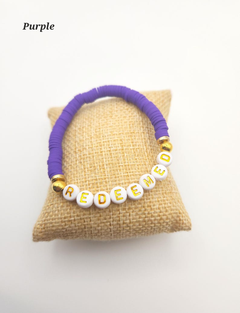 May include: A bracelet featuring purple disc-shaped beads and white spherical beads with the gold-colored text "REDEEMED" printed on them. The bracelet includes two gold-colored spherical accent beads positioned between the purple and white beads.