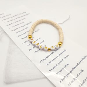 May include: A stretch bracelet made of small, round, beige clay beads featuring white letter beads that spell out PSALM 23 in gold font, accented with small, round, metallic gold beads, resting on a white card printed with the text of Psalm 23.