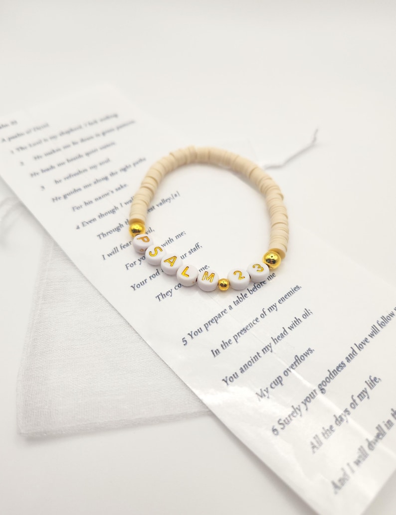 May include: A stretch bracelet made of small, round, beige clay beads featuring white letter beads that spell out PSALM 23 in gold font, accented with small, round, metallic gold beads, resting on a white card printed with the text of Psalm 23.