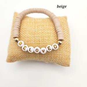 May include: A beige bracelet made of flat circular clay beads featuring white spherical beads with gold lettering that spells out the word BELOVED, accented by two small gold-toned metallic spheres on either side of the lettered beads.