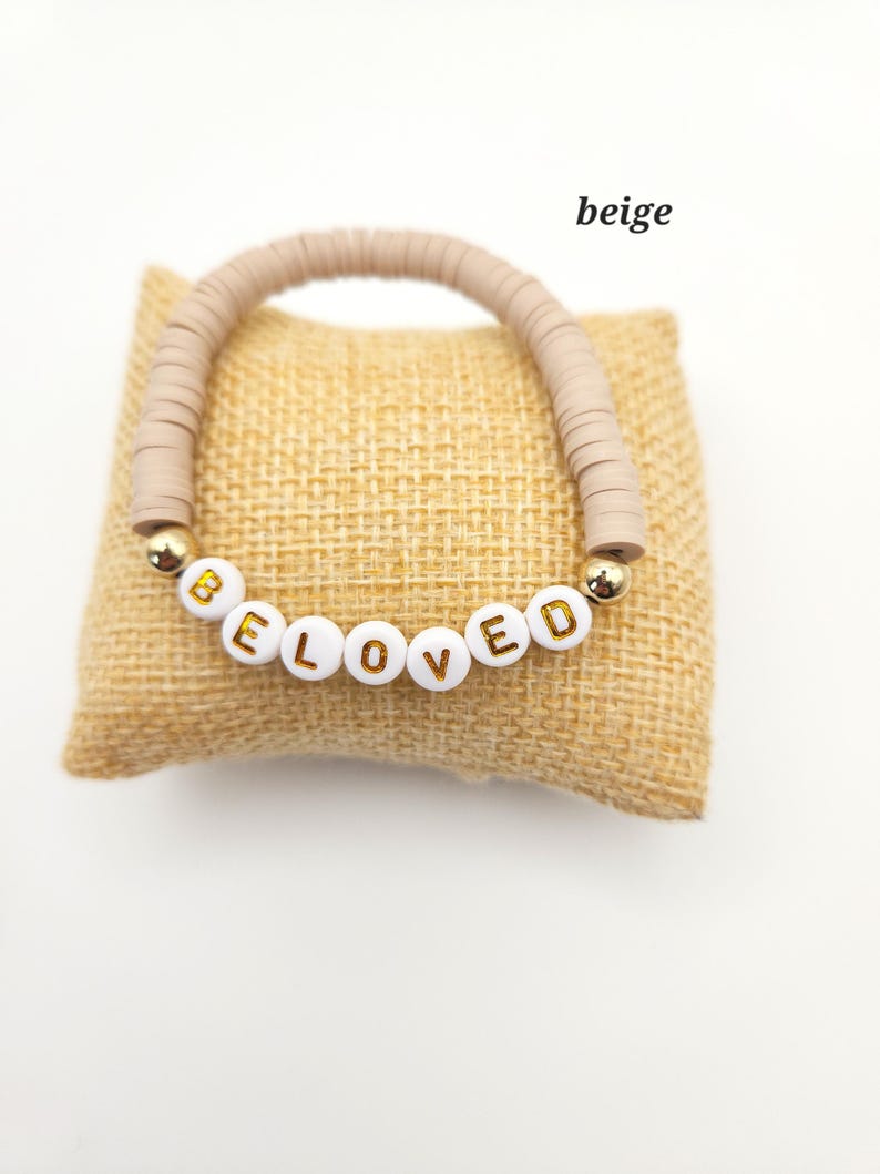 May include: A beige bracelet made of flat circular clay beads featuring white spherical beads with gold lettering that spells out the word BELOVED, accented by two small gold-toned metallic spheres on either side of the lettered beads.