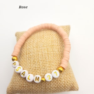 May include: A bracelet made of light pink disc-shaped beads featuring white spherical beads with gold lettering that spells out PSALM 21, interspersed with small gold-colored spherical beads. The word Rose is printed in black text above the bracelet.