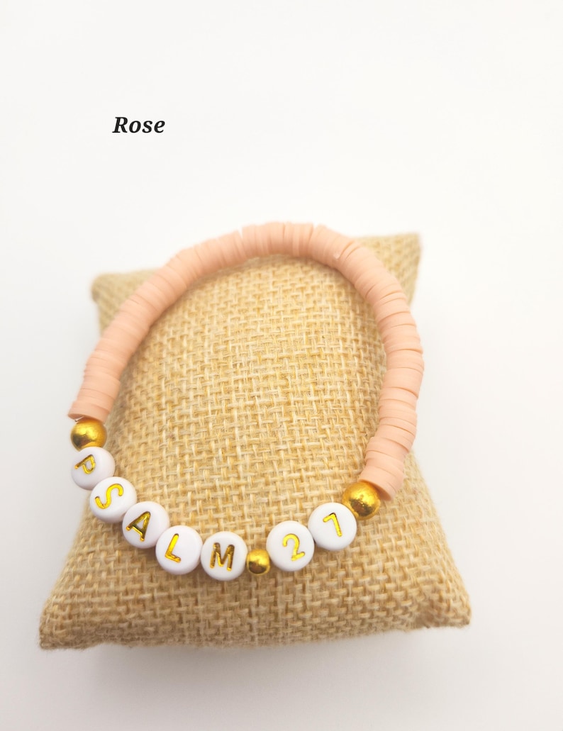 May include: A bracelet made of light pink disc-shaped beads featuring white spherical beads with gold lettering that spells out PSALM 21, interspersed with small gold-colored spherical beads. The word Rose is printed in black text above the bracelet.