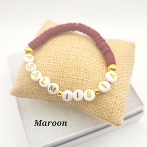 May include: A maroon beaded bracelet featuring white circular beads with gold lettering that spells out PSALM 118 14. The bracelet includes small gold-colored spacer beads interspersed between the maroon and white beads, creating a distinct pattern.