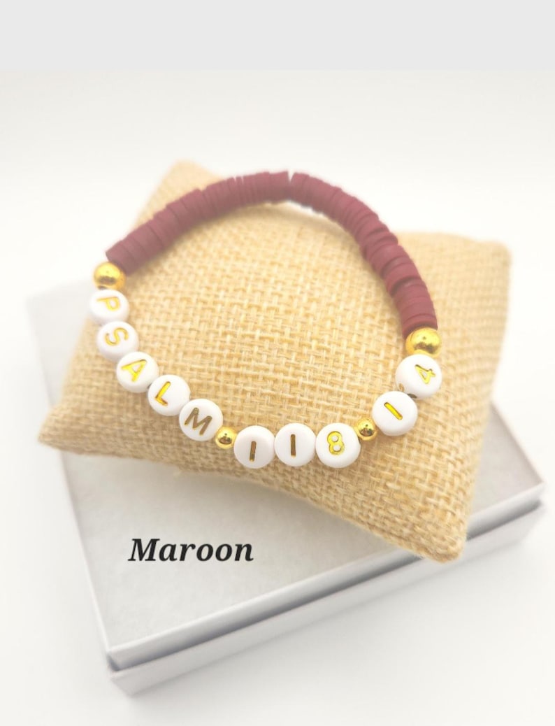 May include: A maroon beaded bracelet featuring white circular beads with gold lettering that spells out PSALM 118 14. The bracelet includes small gold-colored spacer beads interspersed between the maroon and white beads, creating a distinct pattern.