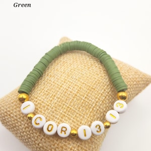May include: A stretch bracelet featuring olive green disc-shaped beads and white spherical beads with gold lettering. The white beads spell out I COR 13 13, interspersed with small gold-colored metallic spacer beads throughout the design.