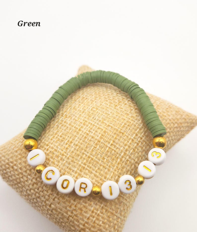 May include: A stretch bracelet featuring olive green disc-shaped beads and white spherical beads with gold lettering. The white beads spell out I COR 13 13, interspersed with small gold-colored metallic spacer beads throughout the design.