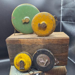 Peut inclure: Quatre rubans à mesurer ronds de style vintage avec des boîtiers en métal vert, jaune et noir. Deux rubans plus grands sont placés en haut, tandis que deux plus petits sont en bas. Chacun possède une manivelle centrale et des détails en métal.