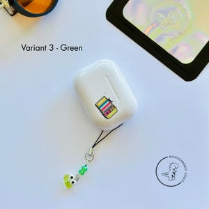 May include: A white AirPod case featuring a colourful retro TV sticker. The case has a black lanyard with green beads and a lime green charm. The text "Variant 3 - Green" is visible.