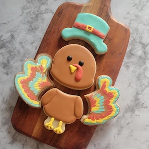 May include: A set of four Thanksgiving-themed sugar cookies decorated with colourful icing. The cookies are shaped like a turkey with a hat, a turkey body, and two turkey wings.