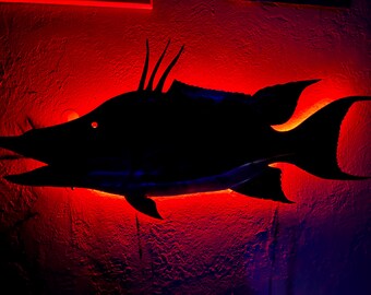 Puede incluir: Decoración de pared con forma de pez negro con un resplandor rojo y naranja. La silueta del pez destaca sobre el fondo rojo, con detalles como aletas y púas visibles. La iluminación crea un efecto dramático.