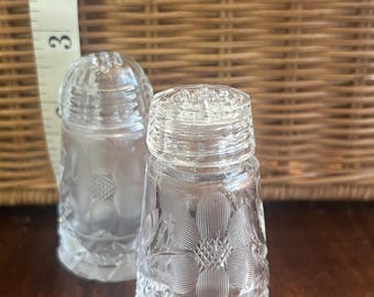 May include: Two clear glass salt and pepper shakers with a floral design. The shakers are on a wooden surface.