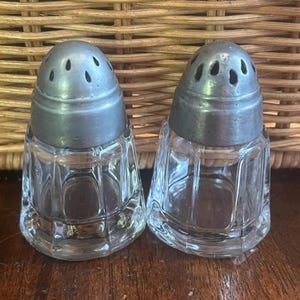 May include: A pair of vintage glass salt and pepper shakers with silver tops. The shakers have a clear glass body with a faceted design and a silver top with holes for dispensing salt and pepper.