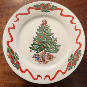 May include: A white ceramic Christmas plate with a green and red ribbon border. The center of the plate features a green Christmas tree decorated with ornaments, a toy soldier, a rocking horse, and a teddy bear. There are presents at the base of the tree.
