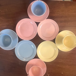 May include: A collection of pastel-colored teacups and saucers arranged on a dark wooden surface. The set includes pink, blue, and yellow cups and saucers. The arrangement suggests a tea party or a display of vintage tableware.