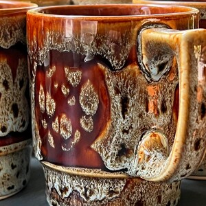 May include: Set of ceramic mugs with a unique glaze. The mugs feature a rich brown and tan color palette with a textured, dripped glaze effect. The handles are a lighter shade of brown. The mugs are ideal for coffee or tea.