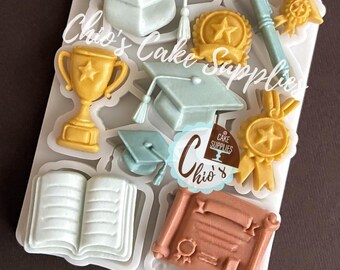 May include: A white silicone mold filled with graduation-themed shapes. Includes mortarboards, a trophy, a pen, a book, a scroll, and medals in various colors. The text "Chio's Cake Supplies" is visible.