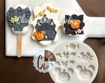 May include: A white silicone mold featuring cavities for Halloween-themed shapes including bats, a spider, and pumpkins with mouse-ear silhouettes. Beside the mold are three decorated cookies featuring a candelabra, a haunted house, and a framed pumpkin.