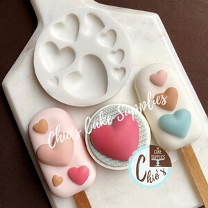 May include: A silicone mold with multiple heart shapes, a round cake pop with a pink heart design, and two popsicle-shaped cake pops with pink, brown, and blue hearts. The cake pops are on wooden sticks. The text "Chio's Cake Supplies" is visible on the image.