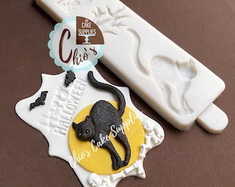 May include: A white silicone mold with a cat silhouette and a black cat cake decoration. The cake decoration features a black cat on a yellow moon, surrounded by bones and bats, with "Happy Halloween" text.