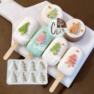 May include: A white silicone mold featuring eight distinct Christmas tree designs alongside four cake popsicles and one round cake treat, all decorated with small edible green, pink, or tan trees and gold sprinkles, labeled Chio's Cake Supplies.