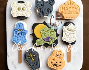 May include: A collection of Halloween-themed cookies on sticks, including a skull, black cat, owl, witch, ghost, coffin, and jack-o'-lantern. The cookies are decorated with various colors and designs, and the words "HAPPY HALLOWEEN" are visible on one cookie.