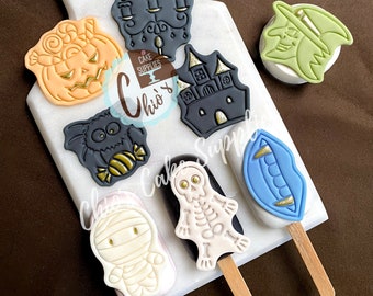 May include: A collection of Halloween-themed cake pops on a white cutting board. The pops feature designs like a pumpkin, a bat with candy, a mummy, a skeleton, a witch, a haunted house, and a candelabra. Each pop is on a wooden stick.
