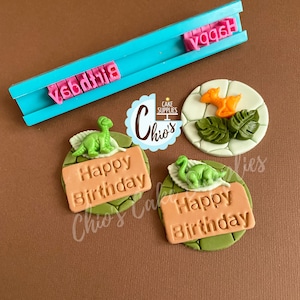 May include: Three green and brown fondant cake toppers with a dinosaur and the words "Happy Birthday" on a brown rectangle. The toppers are on a white background. There is also a blue and pink fondant stamp with the words "Happy Birthday" on it.