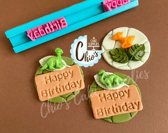 Puede incluir: Tres adornos de pastel de fondant verde y marrón con un dinosaurio y las palabras "Feliz cumpleaños" en un rectángulo marrón. Los adornos están sobre un fondo blanco. También hay un sello de fondant azul y rosa con las palabras "Feliz cumpleaños" en él.