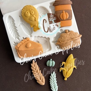 May include: A white silicone mould featuring autumn-themed shapes: a turkey, pumpkin spice latte, pie, and corn. Also includes wheat stalks and a small pumpkin. The text "Chio's Cake Supplies" is visible.
