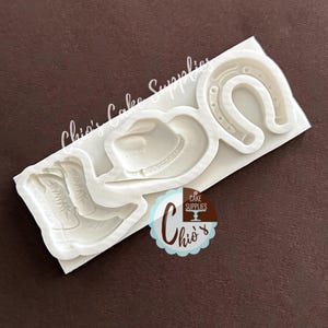 Könnte beinhalten: Eine weiße Silikonform mit geprägten Motiven eines Cowboyhuts, eines Paares Westernstiefel und eines Hufeisens. Die Form enthält den Text Chio's Cake Supplies auf der Oberfläche sowie ein kreisförmiges Logo mit dem Text Chio's Cake Supplies.
