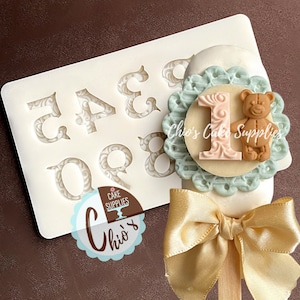 Peut inclure: Un moule en silicone blanc avec les chiffres de 0 à 9 dans une police décorative. Le moule est utilisé pour créer des cake pops ou d'autres décorations comestibles. Un cake pop avec un cercle à motifs bleu et blanc, un numéro 1 rose et un ours en peluche brun est présenté à côté du moule.