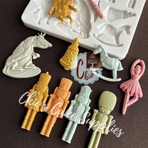 Puede incluir: Una colección de coloridas decoraciones para pasteles moldeadas en varias formas, que incluyen cascanueces, una bailarina, un caballito balancín y una campana. Las decoraciones son en tonos de amarillo, naranja, verde y rosa. También se ve un molde de silicona blanco con formas similares.