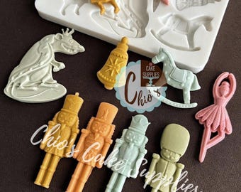Könnte beinhalten: Eine Sammlung farbenfroher, geformter Kuchendekorationen in verschiedenen Formen, darunter Nussknacker, eine Ballerina, ein Schaukelpferd und eine Glocke. Die Dekorationen sind in Gelb-, Orange-, Grün- und Rosatönen gehalten. Eine weiße Silikonform mit ähnlichen Formen ist ebenfalls sichtbar.