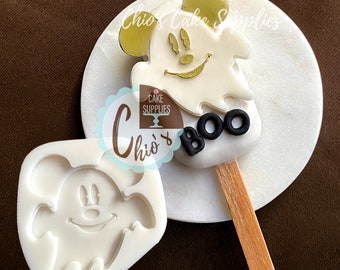 May include: A white Mickey Mouse ghost-shaped cake pop with gold detailing and the word "BOO" in black. It's on a wooden stick and a white marble surface. A silicone mould of the same design is also visible.
