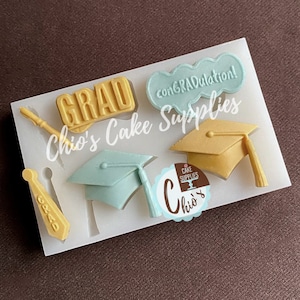 以下が含まれることがあります： 卒業をテーマにしたケーキのデコレーションを作るためのシリコンモールド。モールドには、卒業帽、巻物、ネクタイ、そして「GRAD」と「おめでとうございます！」と書かれた2つの看板が含まれています。