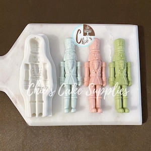 May include: A white marble cutting board holds a white silicone mould and three colourful nutcracker figurines. The figurines are pastel blue, pink, and green. The mould is in the shape of a nutcracker. The text "Chio's Cake Supplies" is visible.