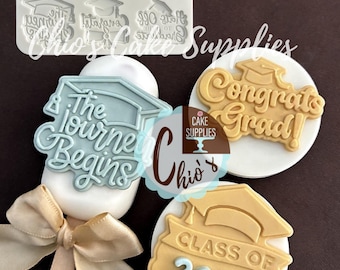May include: A graduation-themed cake decorating set. Includes a white silicone mold with graduation cap designs and edible toppers with phrases like "Congrats Grad!" and "Class of 2026". A light blue "The Journey Begins" cake topper is also visible.