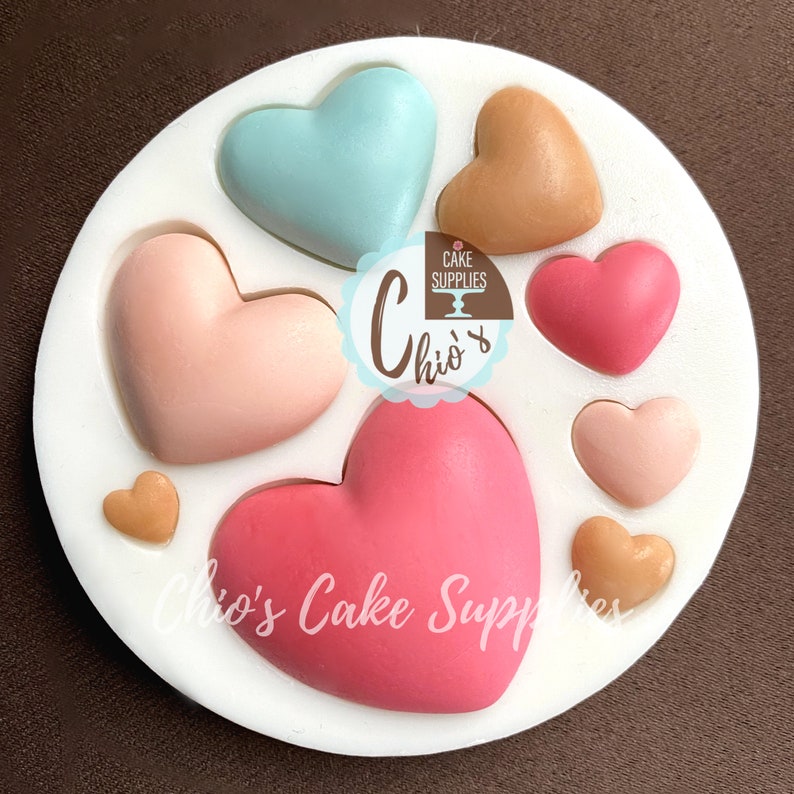 May include: Silicone mold with seven heart-shaped cavities. The mold is white and has a pink, red, blue, and peach heart in each cavity. The mold is labeled "Chio's Cake Supplies".