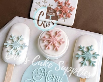 May include: A collection of Christmas-themed cake decorations. Includes white popsicle-shaped treats with blue snowflake designs, a round cake with a pink snowflake, and a blue disc with "Merry Christmas" text. A white mold with snowflake shapes is also present.