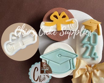 May include: A set of graduation-themed cake pops with a white background. The cake pops are decorated with fondant in the shape of a graduation cap, a diploma, and the year 2024. There is also a white fondant circle with the text "Chio's Cake Supplies" in black.
