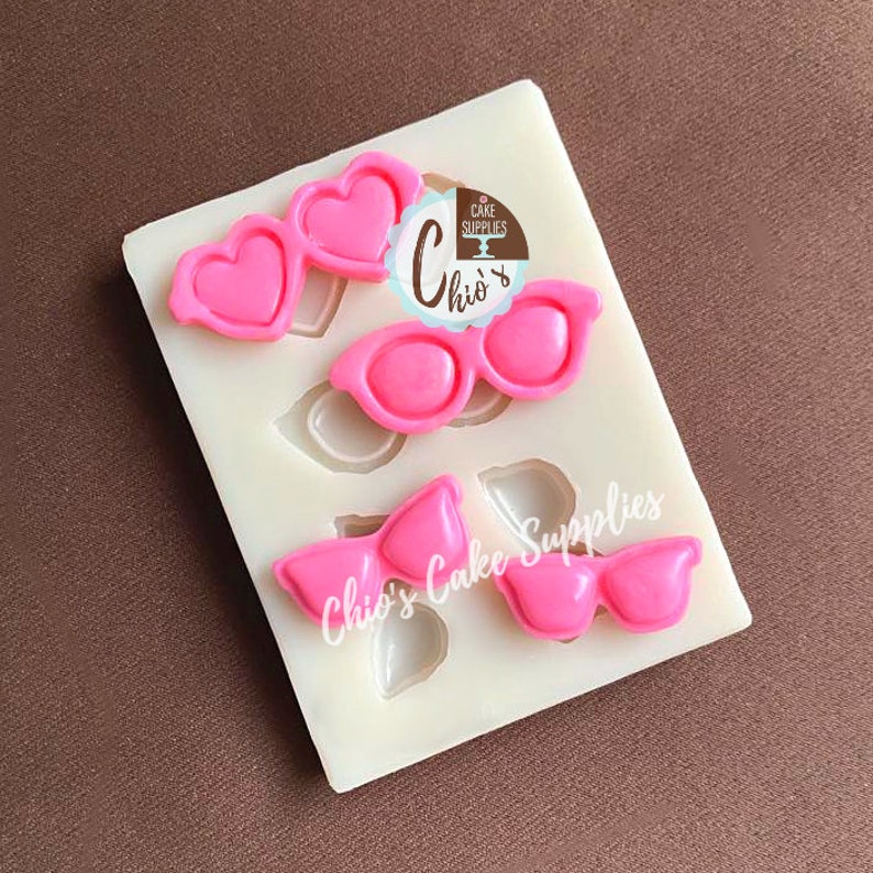 May include: A white silicone mold with three different designs: heart-shaped glasses, round sunglasses, and a bow tie. The designs are filled with bright pink material. The text "Chio's Cake Supplies" is visible.