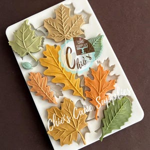 May include: A white silicone mold featuring various autumn leaf designs in shades of green, orange, and brown. The mold includes maple and oak leaf shapes, perfect for creating decorative elements for cakes and other baked goods. The text "Chio's Cake Supplies" is visible.